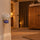 FELIWAY Optimum diffuser plugged into a wall socket in a lamp-lit living room, with a cat resting in a bed on top of a tall cabinet