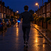 A runner on a dark residential street seen from behind, a blue LED armband glowing evenly along its length on the forearm
