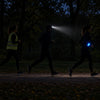 Three figures on a dark path: a hi-vis vest barely visible, a head torch glaring forward, and a glowing LED armband clearly visible from the side