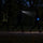 Three figures on a dark path: a hi-vis vest barely visible, a head torch glaring forward, and a glowing LED armband clearly visible from the side
