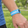 A blue LED armband, switched off, fastened on a man's wrist outdoors in daylight, showing the hook-and-loop strap, the metal loop and the black button.