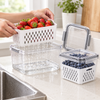Hands lifting a white colander basket of berries out of a clear box in a kitchen, rinse water draining into the box below
