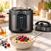 A dark gray Midea multicooker with its lid off beside a bowl of oat porridge topped with raspberries, blueberries and honey, on a marble kitchen counter